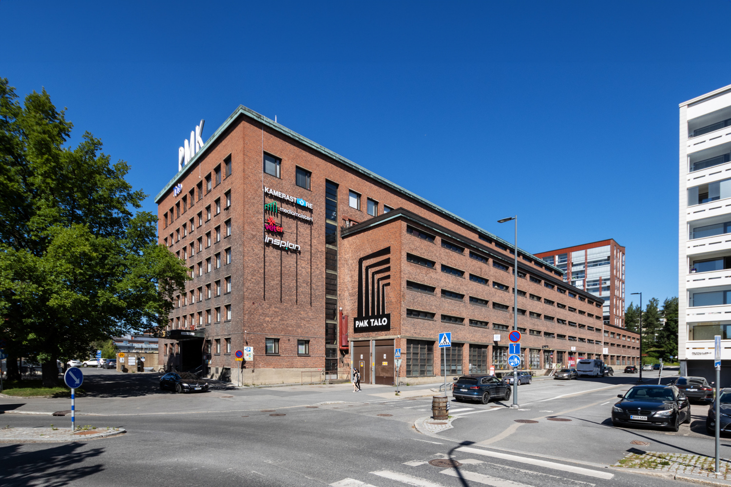 Punatiilinen PMK-talo on 7-kerroksinen teollisuushenkinen toimitilarakennus. Talon edessä on autotie. Kiinteistön logo talon seinässä.